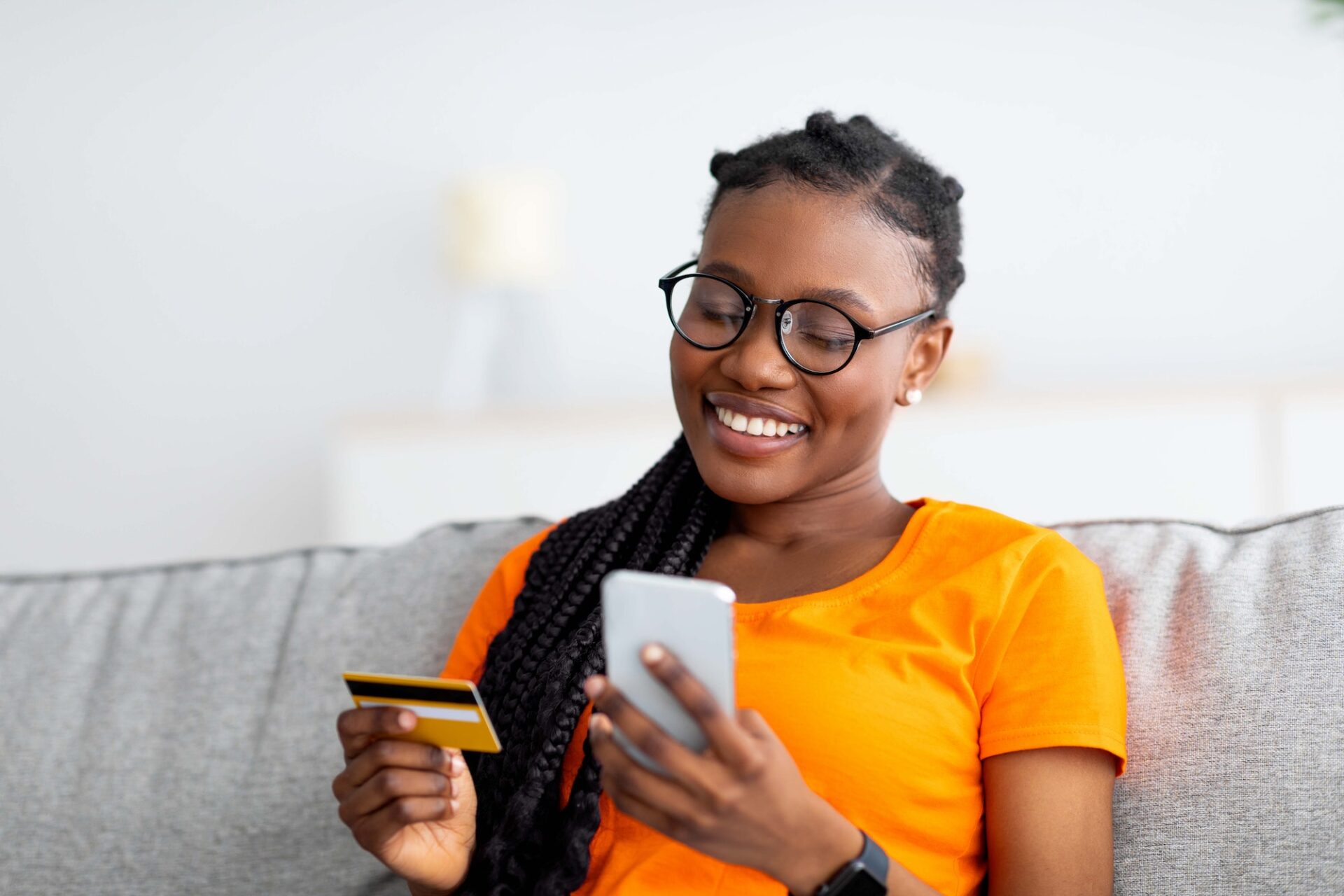 Cheerful young black woman purchasing on web from home, sitting on sofa, using smartphone and credit